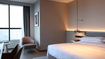 A modern hotel room with a large bed, a desk, a chair, and a view of the city through large windows.