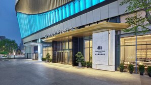 Una entrada de hotel moderna con fachada curva, iluminación azul y un área exterior bien cuidada con plantas en macetas.