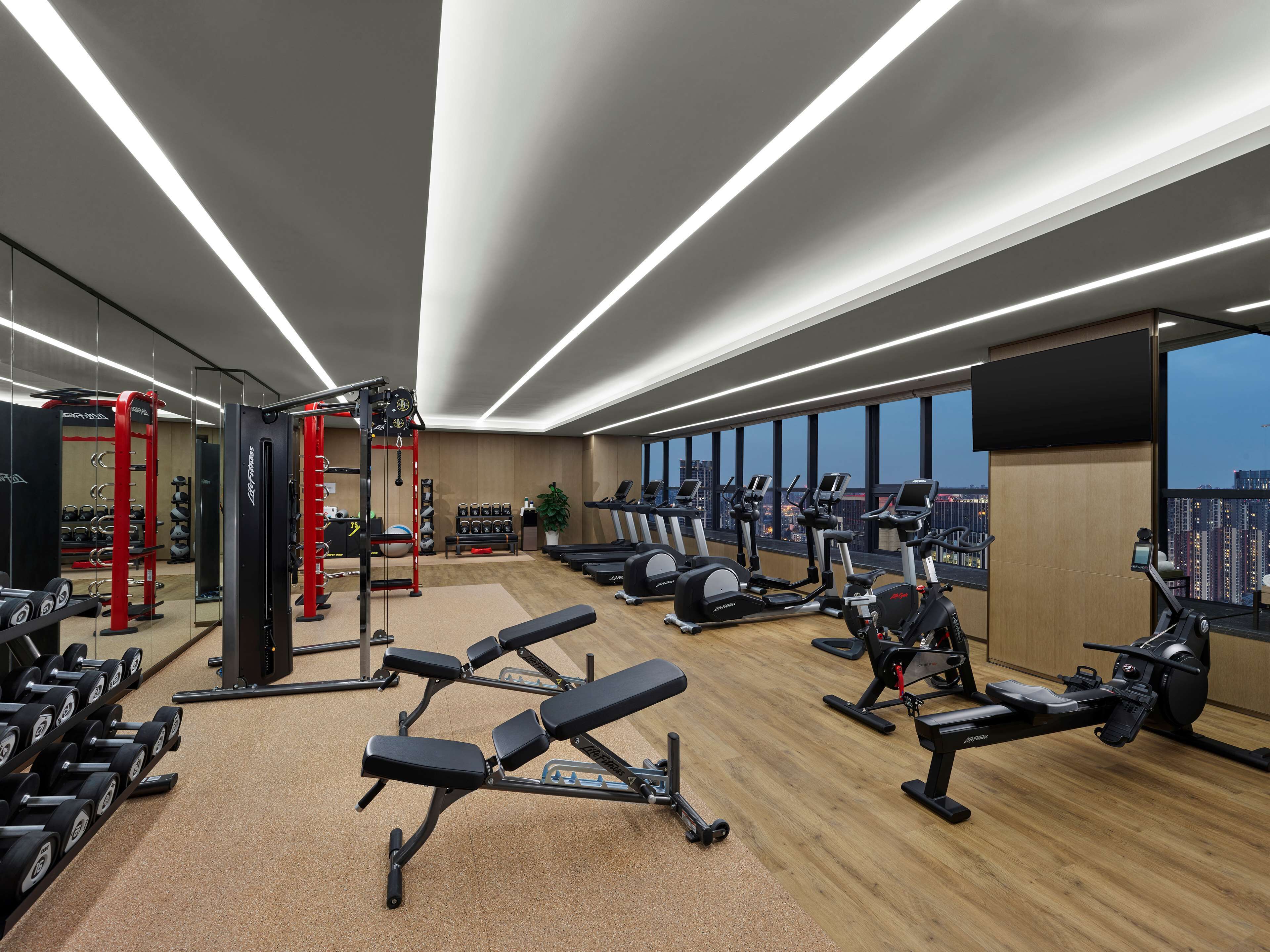 A modern gym with various exercise equipment, a large mirror, and a city view through large windows.
