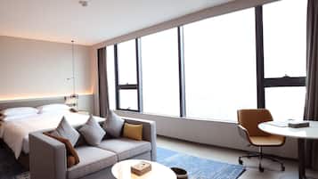 A modern hotel room with a bed, a sofa, a desk, and large windows.