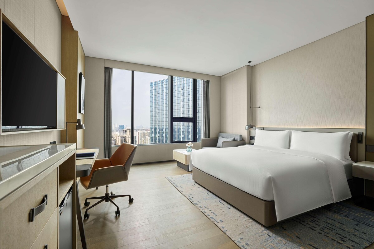 A modern hotel room with a large bed, a desk with a chair, a television, and a view of the cityscape through large windows.