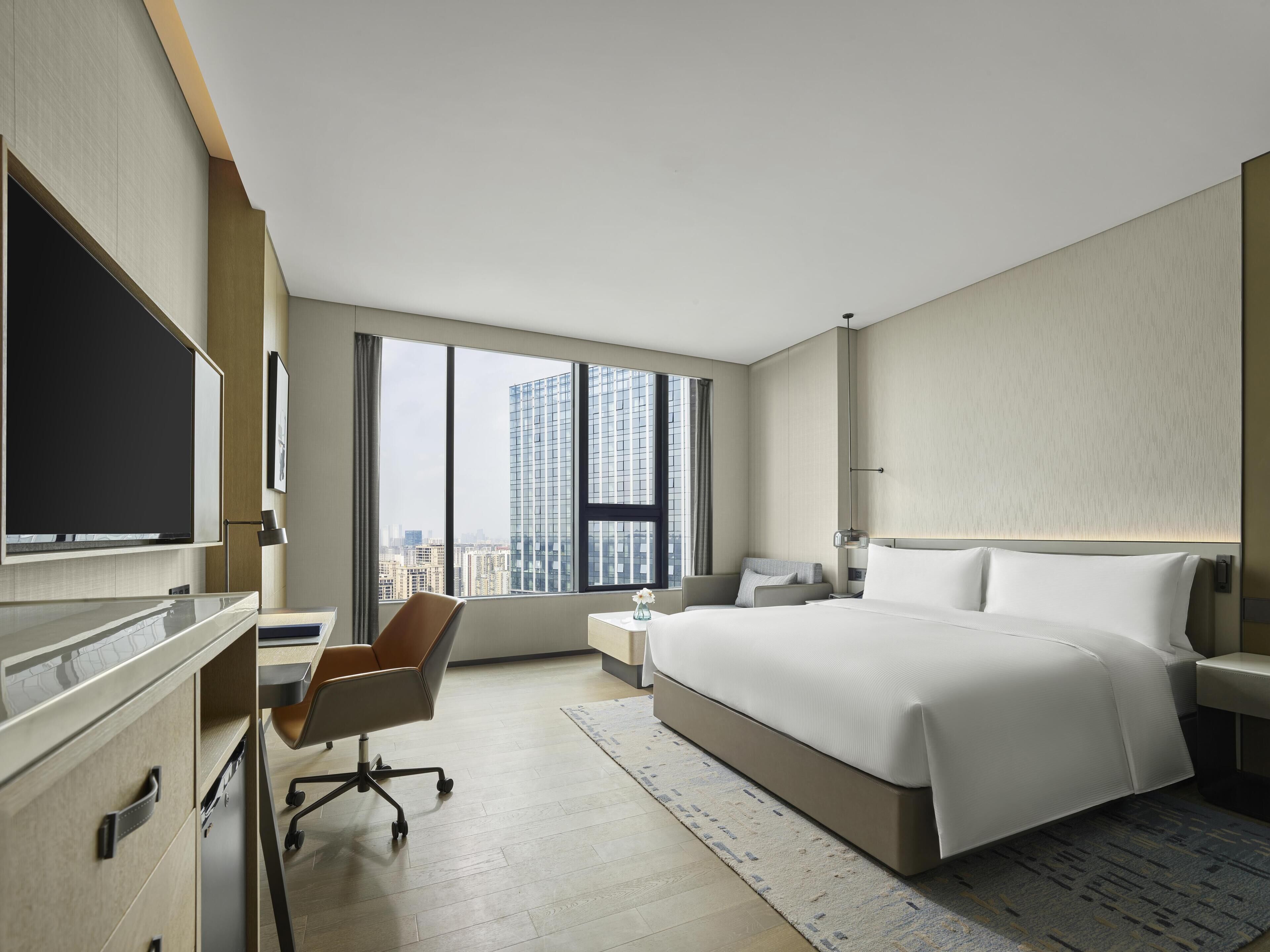 Une chambre d’hôtel moderne dotée d’un grand lit, d’un bureau avec une chaise, d’un téléviseur à écran plat et offrant une vue sur le paysage urbain grâce à de grandes fenêtres.