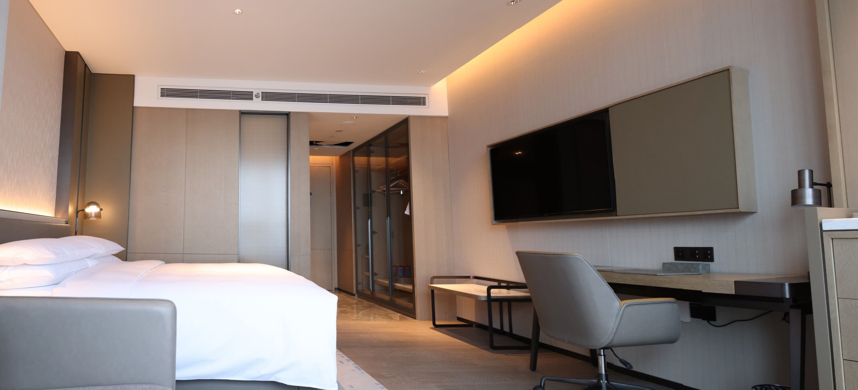 A modern hotel room with a bed, a desk, a chair, a television, and a wardrobe.