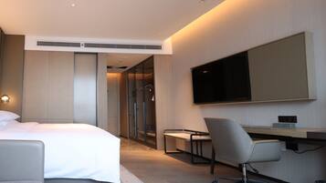 A modern hotel room with a bed, a desk, a chair, a television, and a wardrobe.