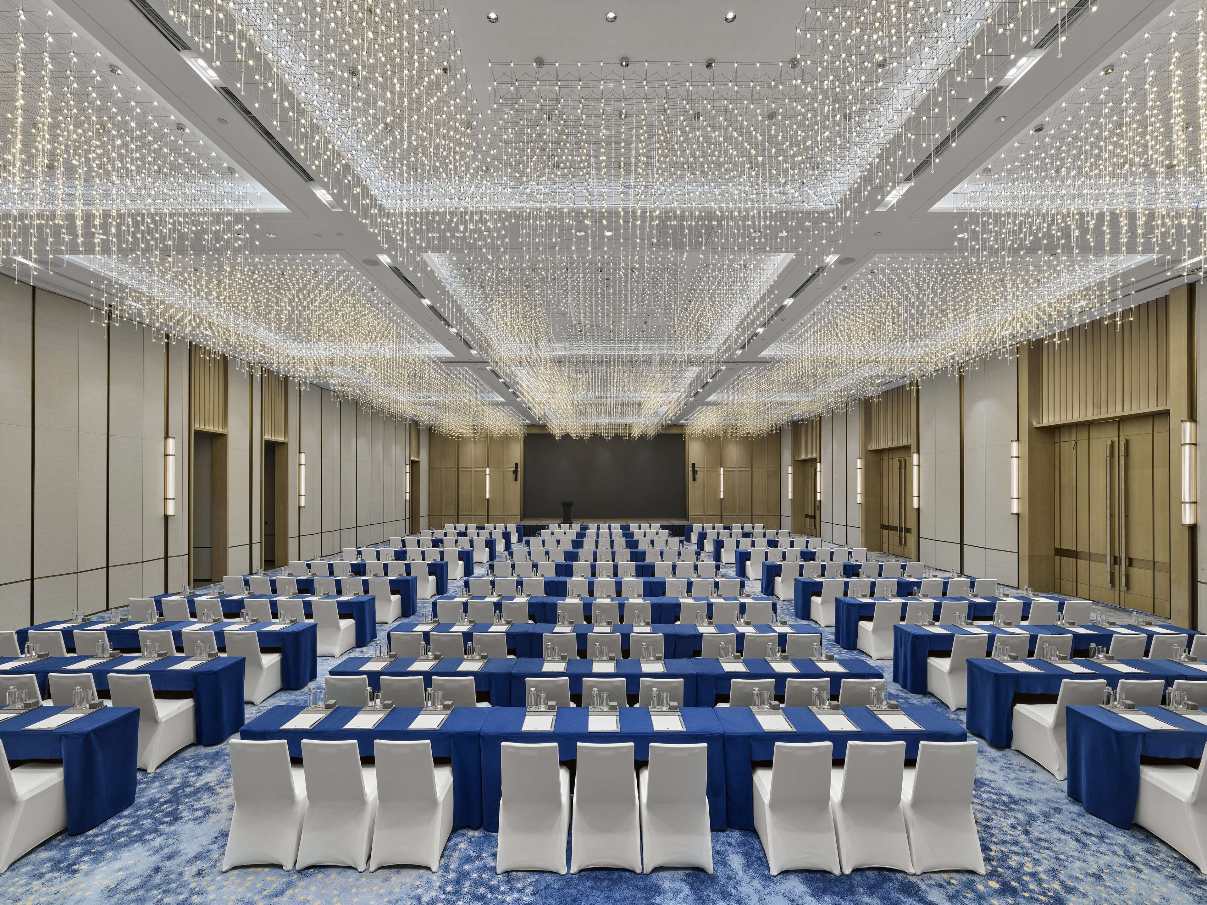 Un amplio salón de conferencias con filas de mesas y sillas, un techo alto con luces colgantes y un escenario al frente.