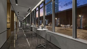 Un pasillo moderno de hotel con mesas y sillas, ventanas amplias y vistas a la ciudad al atardecer.