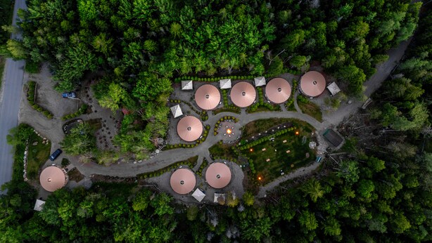 Vista aérea de un camping con tiendas de campaña circulares dispuestas en un semicírculo, rodeado de un bosque denso.