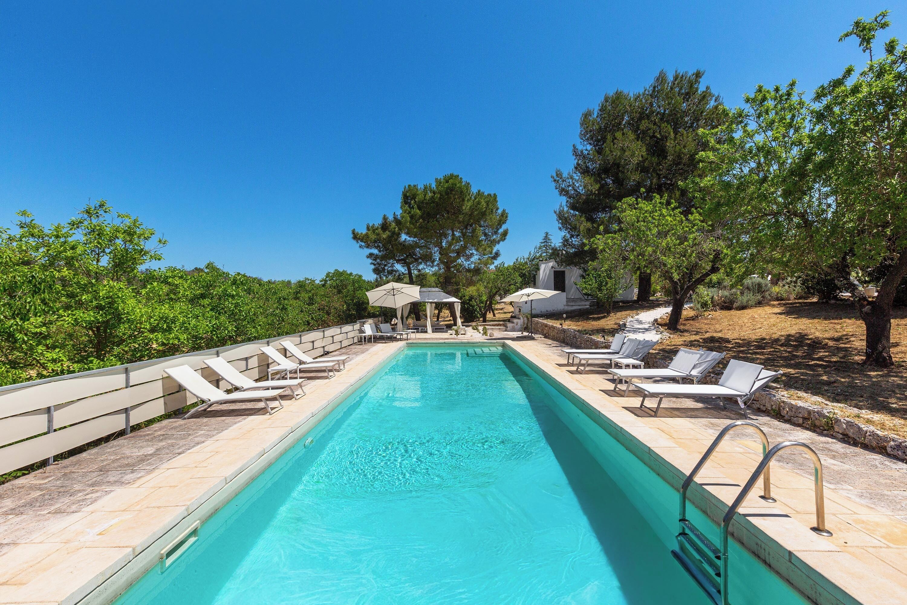 Une piscine rectangulaire à l’eau d’un bleu limpide, entourée de chaises longues et de parasols, avec des arbres et un bâtiment en arrière-plan.