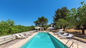 A rectangular swimming pool with a clear blue water surface, surrounded by lounge chairs and umbrellas, with trees and a building in the background.