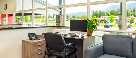 A modern office with a desk, chair, and computer, large windows with a view of a parking lot and mountains.
