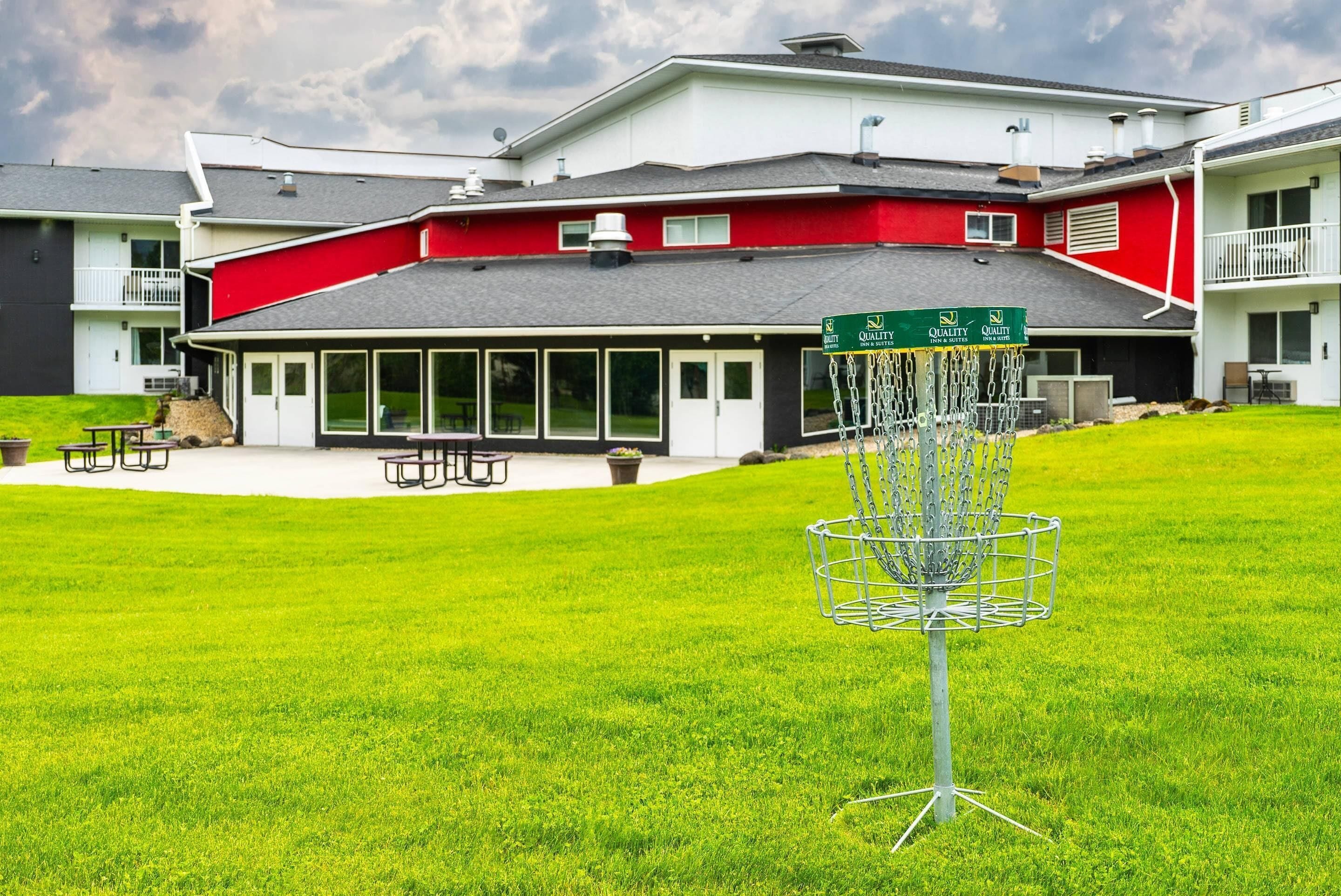 Un campo de disc golf con una canasta metálica en primer plano, un edificio de fachada roja y blanca al fondo y una zona verde con mesas de picnic.