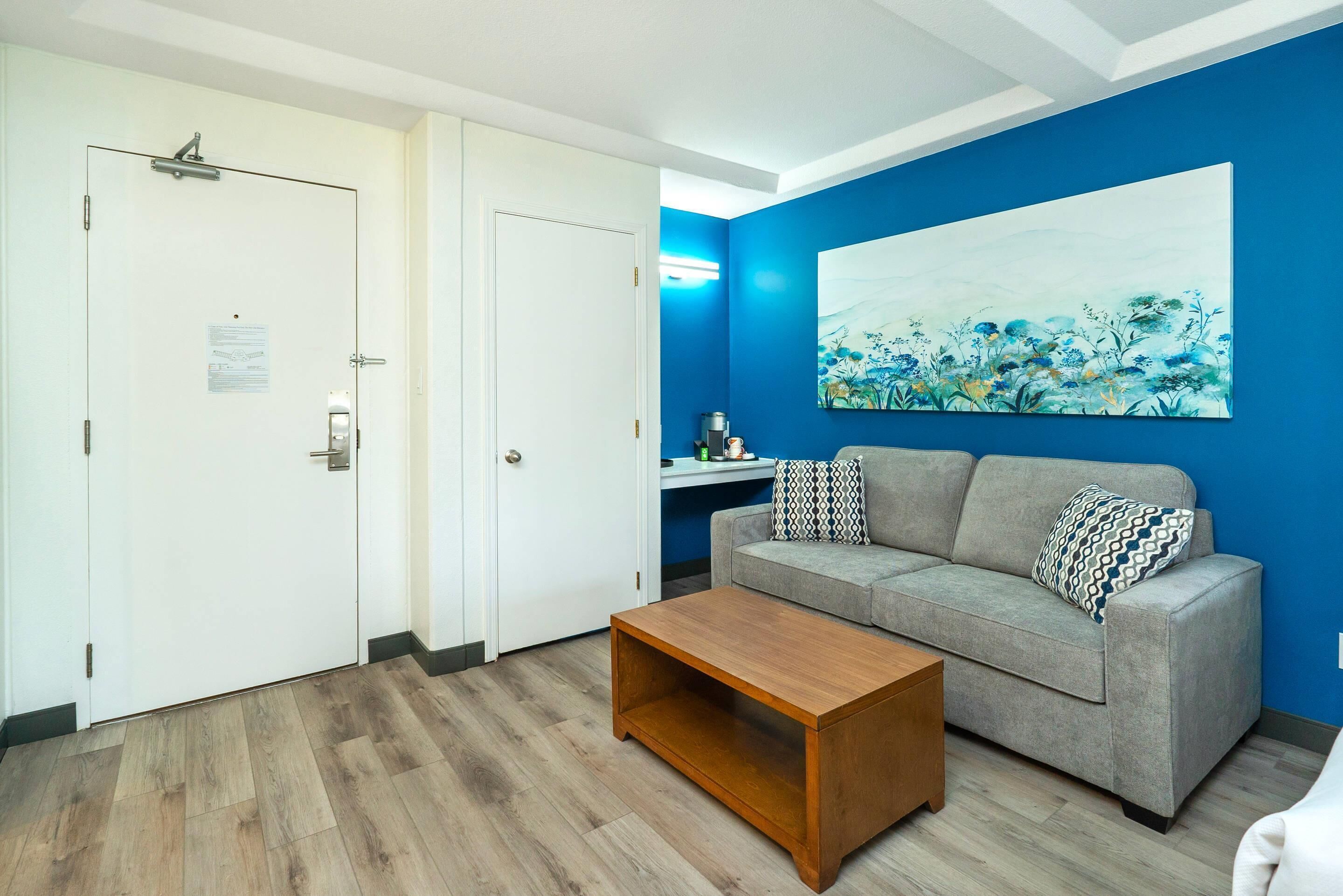 A room with a grey sofa, a wooden coffee table, and a blue wall with a floral painting.