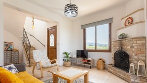 Una sala de estar con sofá, mesa de centro, televisor sobre un soporte de madera, chimenea, escalera y un ventanal con vistas.