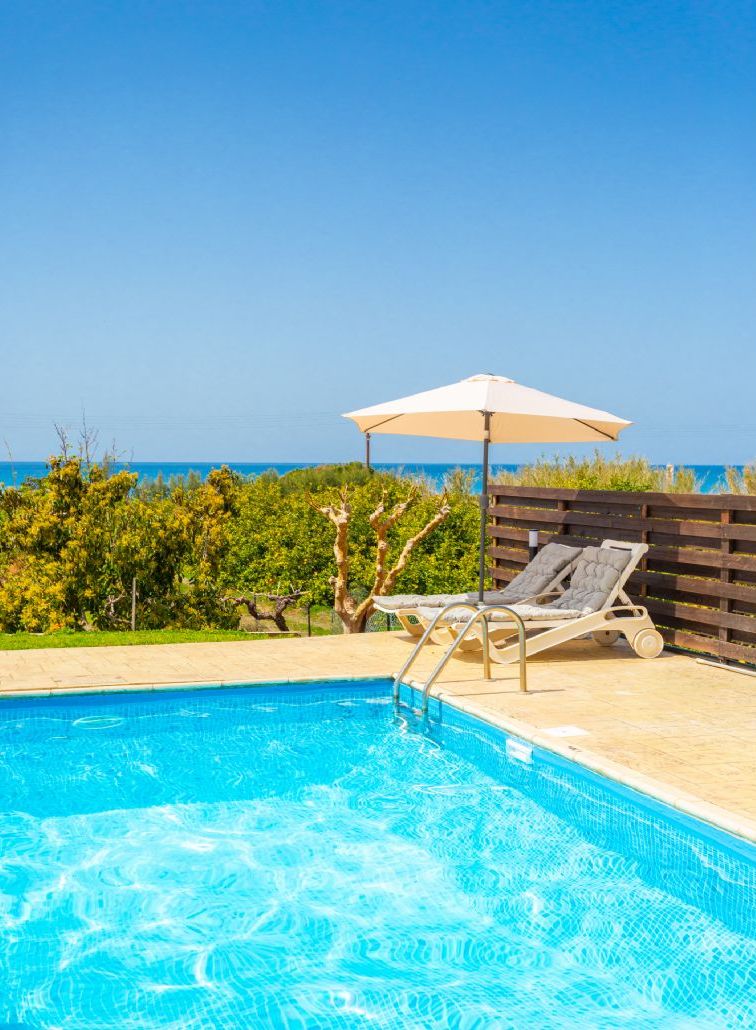 A swimming pool with lounge chairs and an umbrella, surrounded by a wooden fence and greenery.