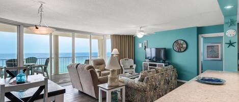 A spacious living area with a large window overlooking the ocean, a comfortable seating arrangement, a television, and a kitchen area.