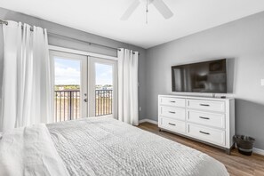 Une chambre avec un grand lit, une commode, une télévision et un balcon avec des rideaux.