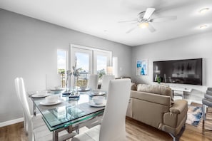 A modern living room with a glass dining table, white chairs, a beige sofa, and a flat-screen TV.