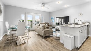 Eine moderne Küche mit Kochinsel, ein Essbereich mit Glastisch und ein Wohnzimmer mit Sofa und Fernseher.