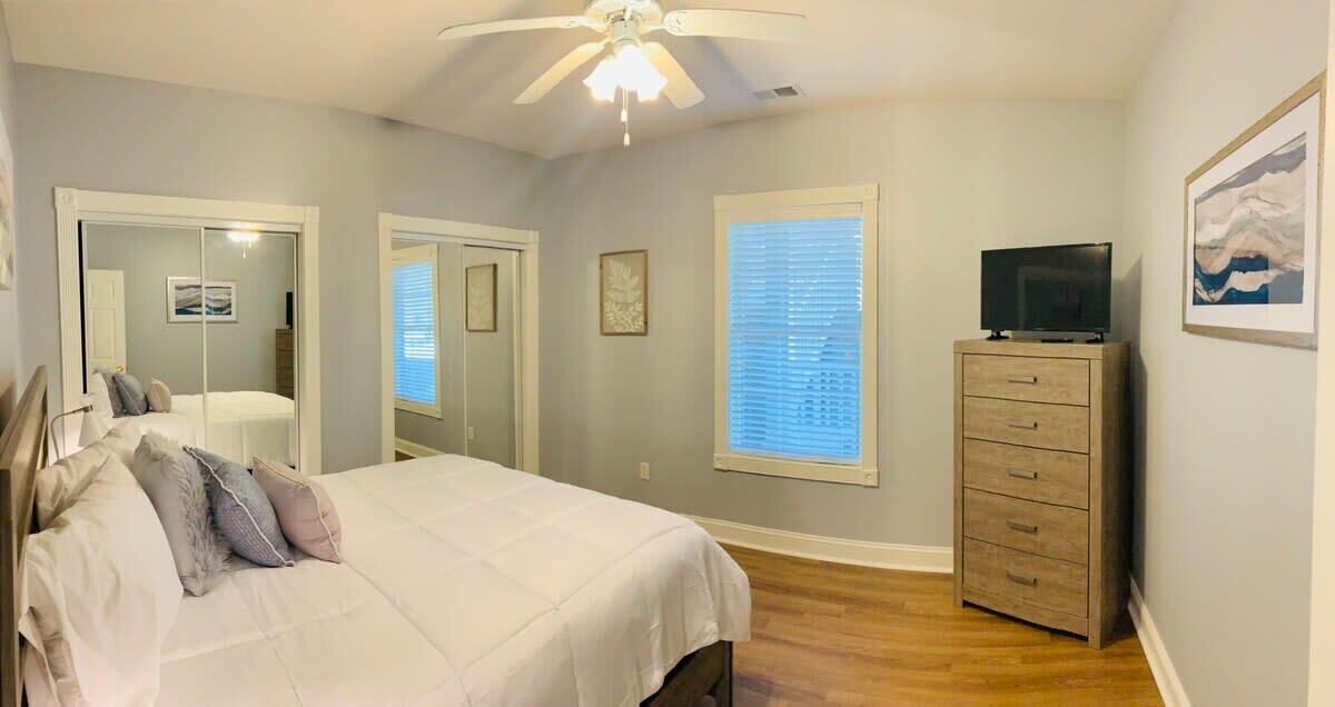 A bedroom with a bed, a ceiling fan, a dresser, a TV, and a window with blinds.