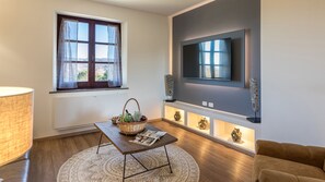 A modern living room with a large flat-screen TV mounted on the wall, a wooden coffee table with a basket and magazine, a patterned rug, and a brown sofa.
