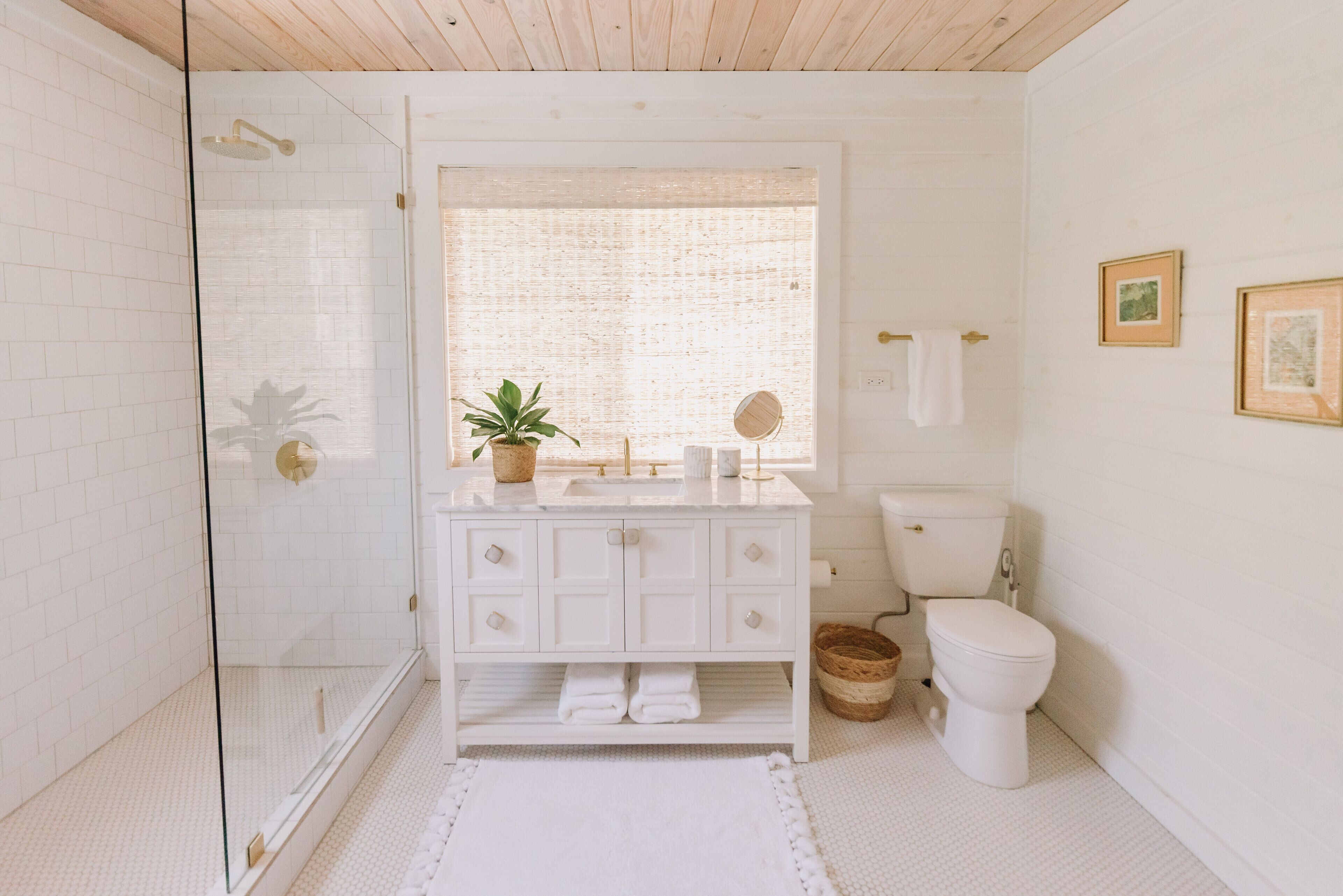 A bathroom with a glass shower enclosure, white tiled walls, a white vanity with drawers, a toilet, and a window with a view of greenery.