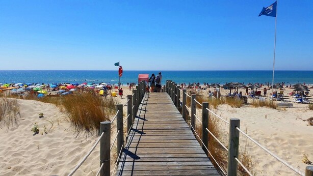 A wooden boardwalk leading to a beach with umbrellas and beachgoers.
