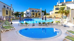 A swimming pool with lounge chairs and umbrellas, surrounded by buildings with balconies and a clear blue sky.