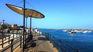 A wooden pier with a metal railing, a large umbrella, and a view of boats on the water.