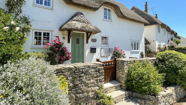 A traditional thatched-roof cottage with a stone wall entrance, a green door, and a signboard. - Cajun Cottage (Wareham)