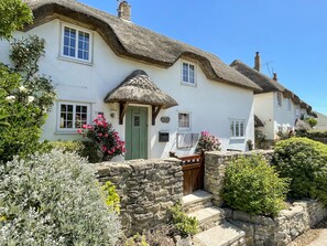 A traditional thatched-roof cottage with a stone wall entrance, a green door, and a signboard. - Cajun Cottage (Wareham)