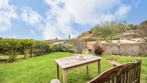 A wooden table with a plate of food and a glass of drink in a grassy outdoor area with a view of a thatched-roof house and hills in the background. - Cajun Cottage (Wareham)
