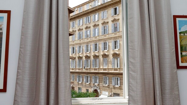 A view through curtains of a multi-story building with a classical architectural style, featuring numerous windows and a central entrance. - 4BNB - Cavour Square Apartment (Rome)