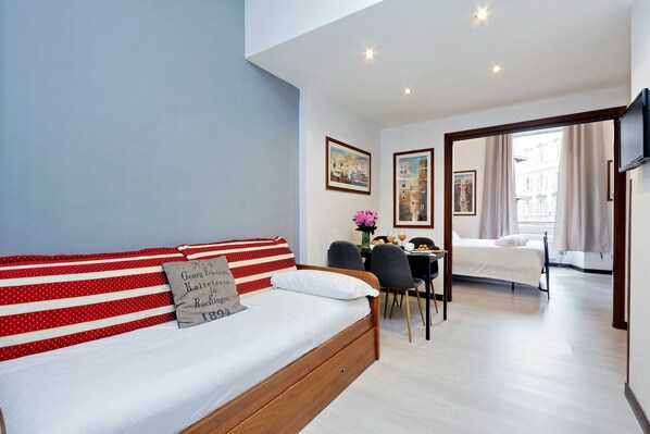 Quarto de hotel moderno com uma cama de madeira, um sofá riscado em vermelho e branco, um espaço de refeições com mesa e cadeiras, e uma TV fixada na parede.