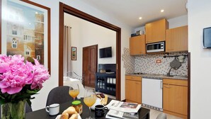 A modern kitchen with wooden cabinets, a microwave, and a sink. There is a dining table with a vase of flowers, a newspaper, and a glass of orange juice. - 4BNB - Cavour Square Apartment (Rome)