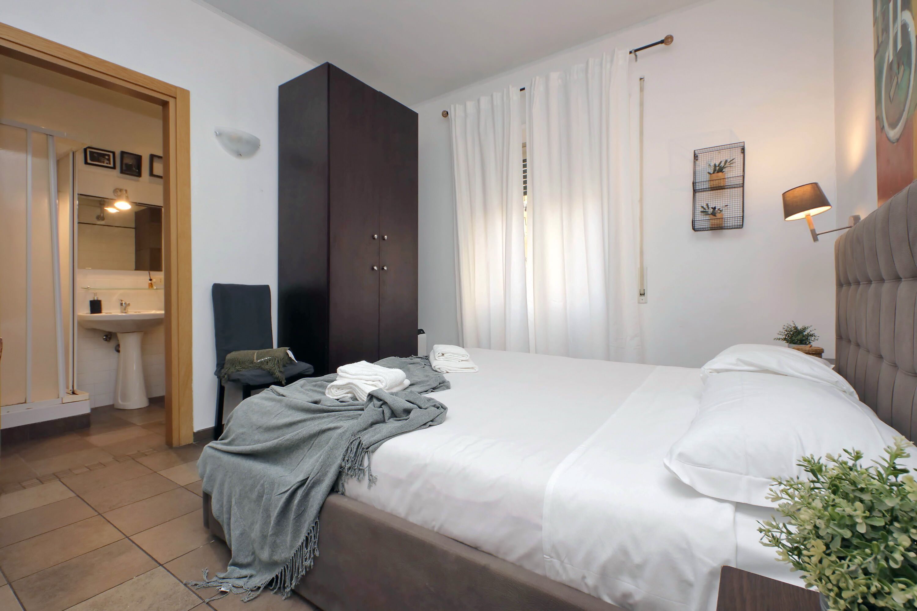 Un dormitorio con cama, armario, mesita de noche, lámpara y una planta pequeña.
