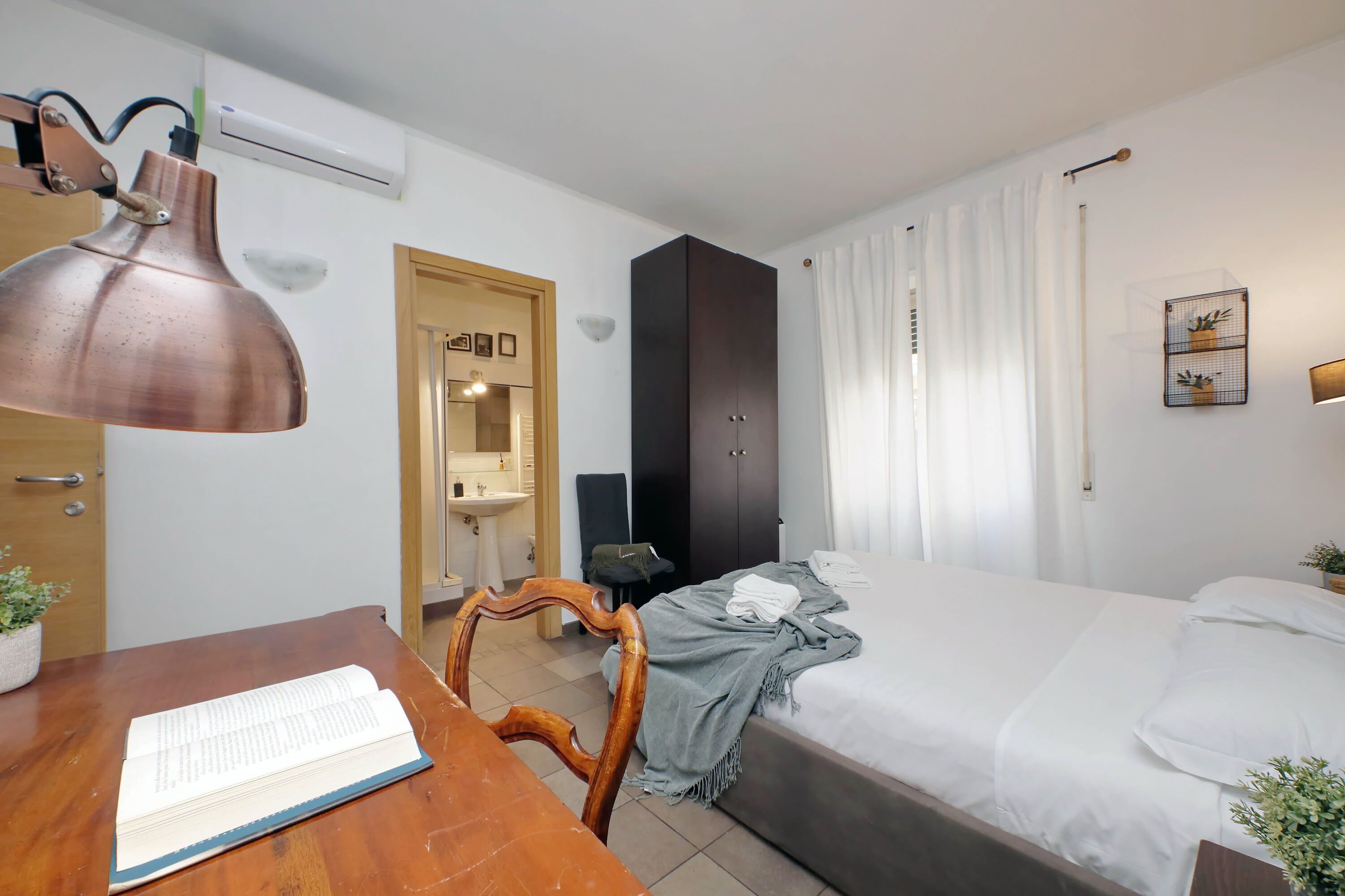 A hotel room with a wooden desk, a bed with white bedding, a grey blanket, and a wall-mounted air conditioner.