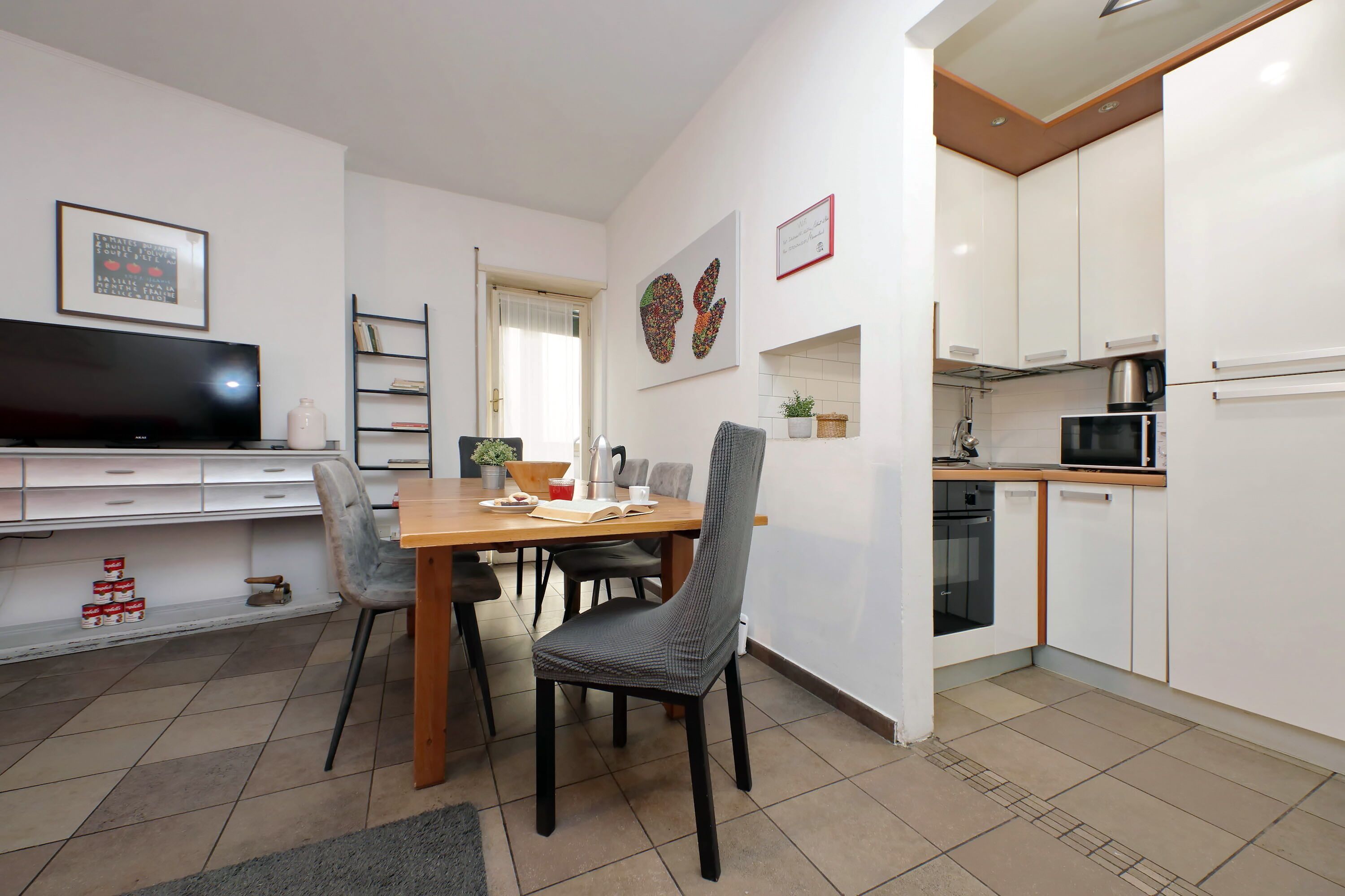 Een moderne keuken met witte kastjes, een magnetron en een koffiezetapparaat. Er is een eethoek met een houten tafel en stoelen.