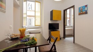 Un comedor con una mesa dispuesta para dos personas, un ramo de flores y un plato de pan. Se observa un televisor sobre un mueble bajo y una ventana con persianas.