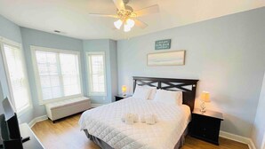 Une chambre à coucher comprenant un lit, des tables de chevet, un ventilateur de plafond et une fenêtre avec des stores.