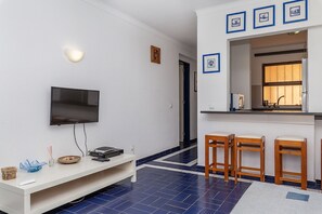 A modern living room with a flat-screen TV, a white entertainment unit, and a kitchen area with bar stools.