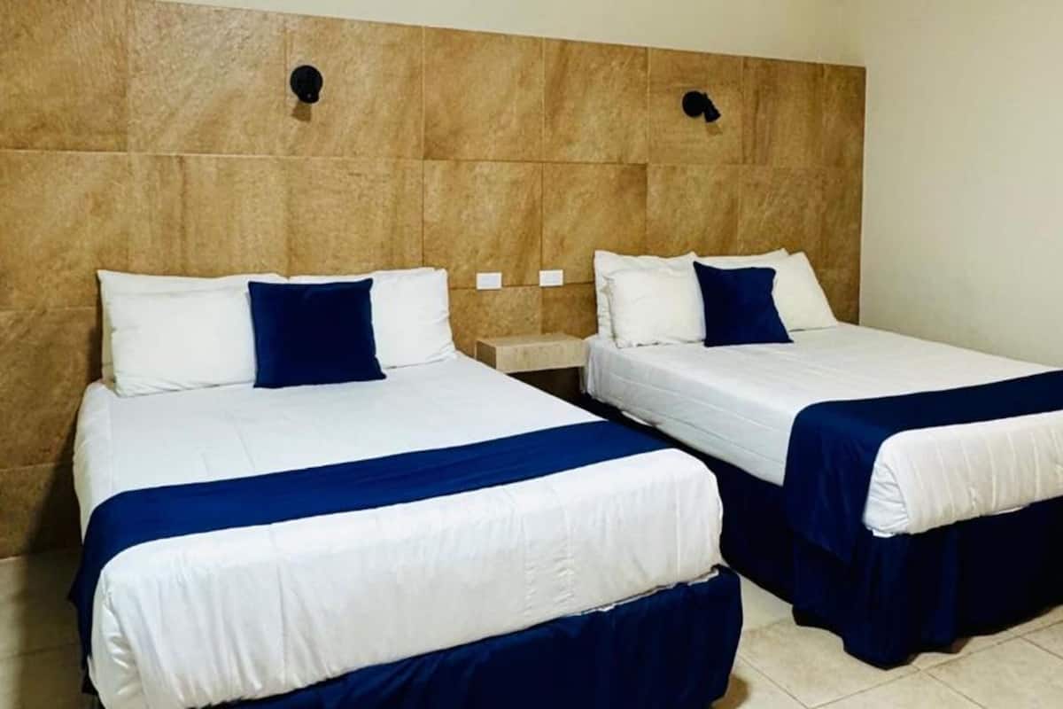 A hotel room with two beds, each with white linens and blue accents, a wooden headboard, and a tiled floor.