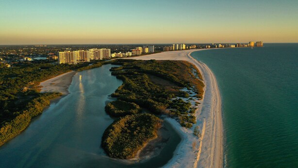 An aerial view of a coastal city with a wide beach, dense greenery, and high-rise buildings in the background. - ANGLERS COVE - WELCOME TO "ISLAND TIME" AT MARCO ISLAND (Marco Island)