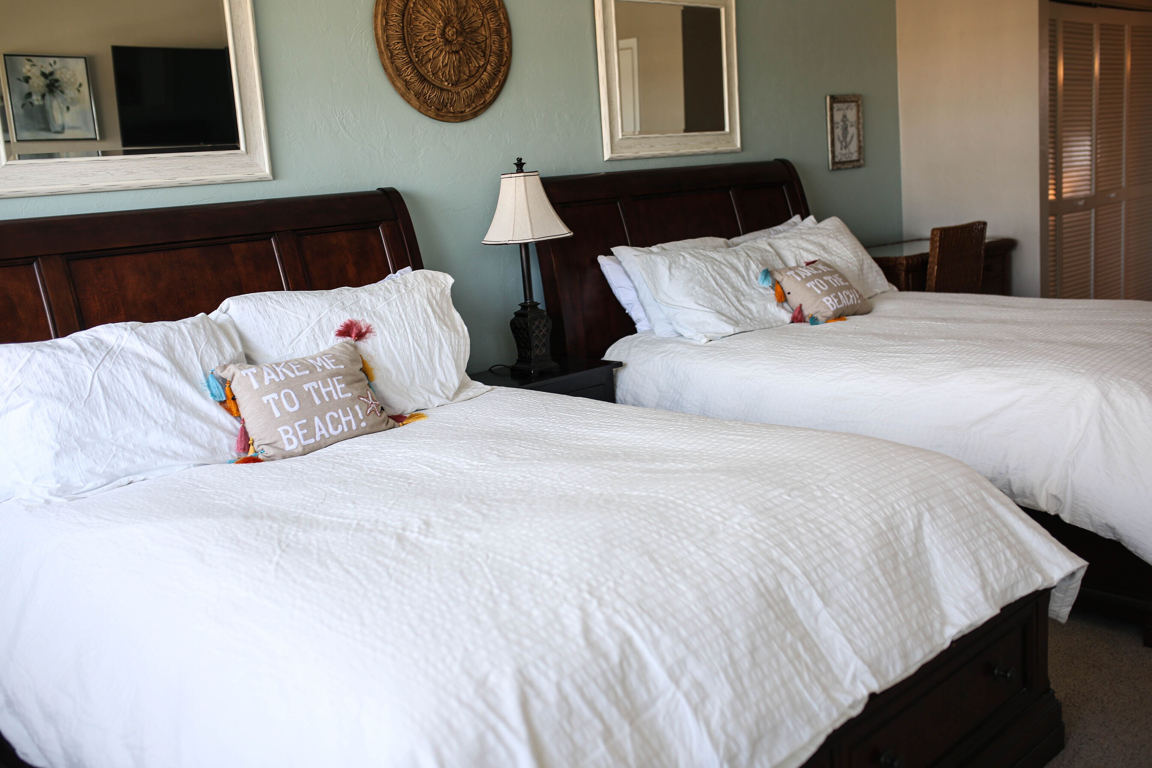 Two neatly made beds with white linens and decorative pillows, a wooden headboard, a bedside lamp, and a wall-mounted mirror.