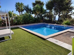 Et rektangulært swimmingpool med flisekant, omgivet af en velplejet græsplæne og forskellige tropiske planter.