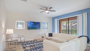 Ein Wohnzimmer mit einem weißen Sofa, einem Couchtisch, einer Lampe und einem Deckenventilator. Zwei Fenster mit Vorhängen, ein Bild an der Wand und ein gemustertes Teppich sind ebenfalls vorhanden.