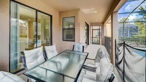 Eine sonnendurchflutete Terrasse mit einem Glastisch und weißen, gepolsterten Sitzgelegenheiten, mit Blick auf einen Garten und ein Gebäude.