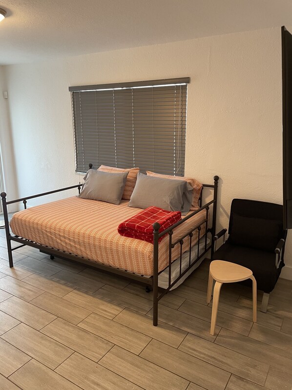 Un lit simple avec un cadre en métal, un petit tabouret et une fenêtre avec des stores.