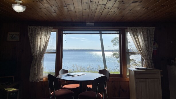 Interior de uma cabana de madeira com uma mesa redonda e cadeiras, um ventilador de teto e uma janela com vista para a água.