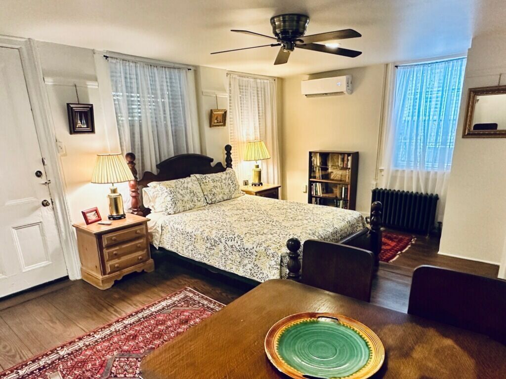A bedroom with a bed, bedside lamps, a wooden nightstand, a red patterned rug, a dining table with a green plate, a ceiling fan, and a bookshelf.