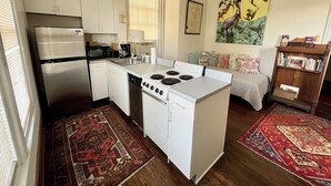 A compact kitchen with a refrigerator, microwave, stove, and dishwasher. There is a ceiling fan, a window with blinds, and a painting on the wall. - George Baldwin House Suite (Savannah)